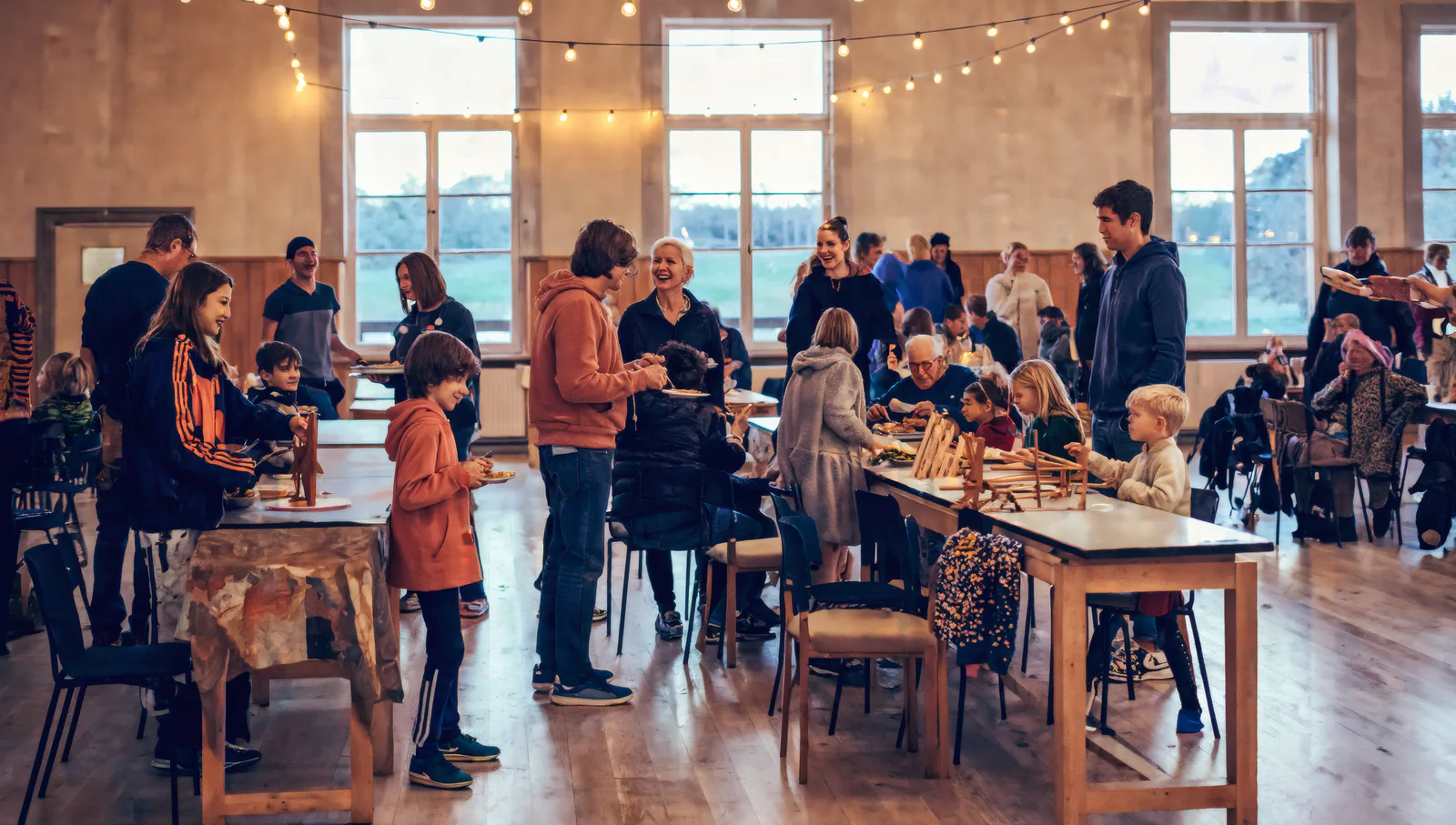 Neighbors of every age sharing a meal at long wooden tables in a warm, light-filled hall