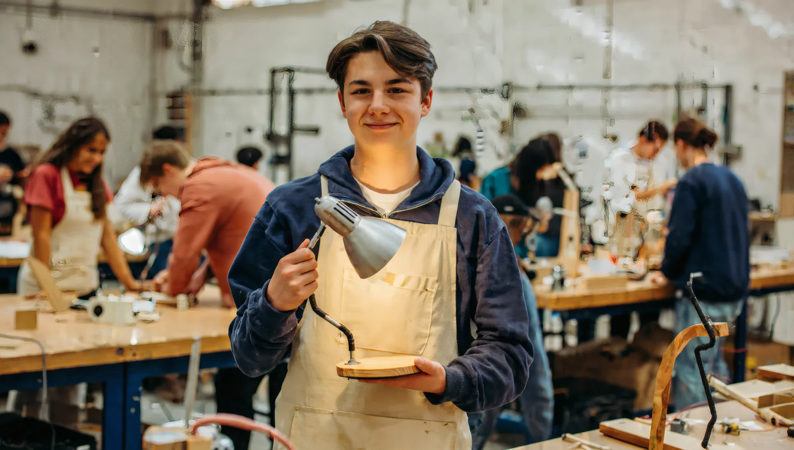 A student in a work apron holding up a desk lamp they built